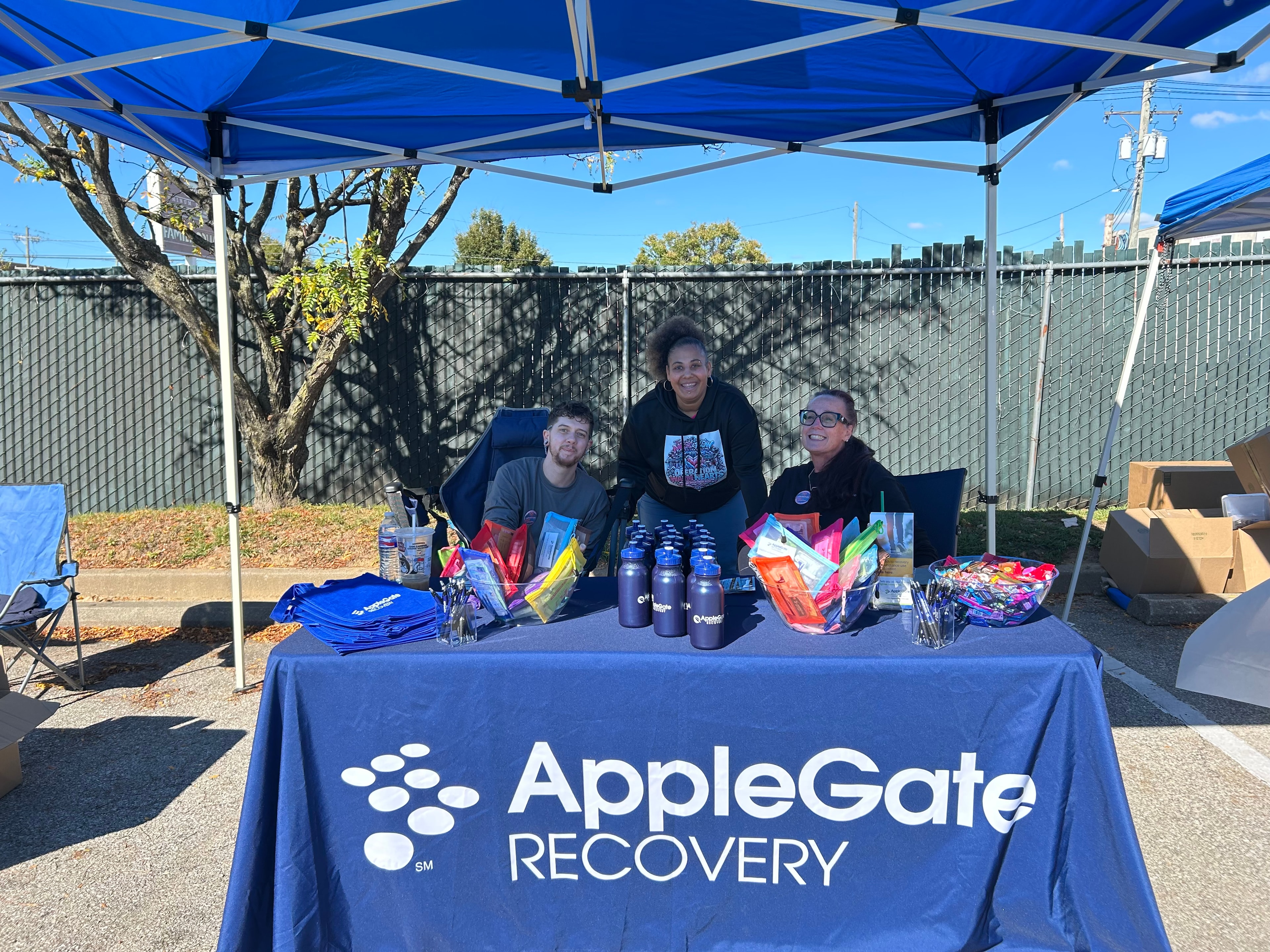 Three people at the AppleGate Recovery booth under a blue canopy at Operation Warm Hearts