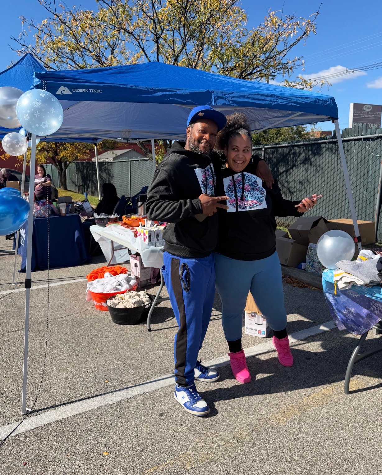 Two people posing together under the blue Ozark Trail tent at Operation Warm Hearts with balloons and supplies in the background