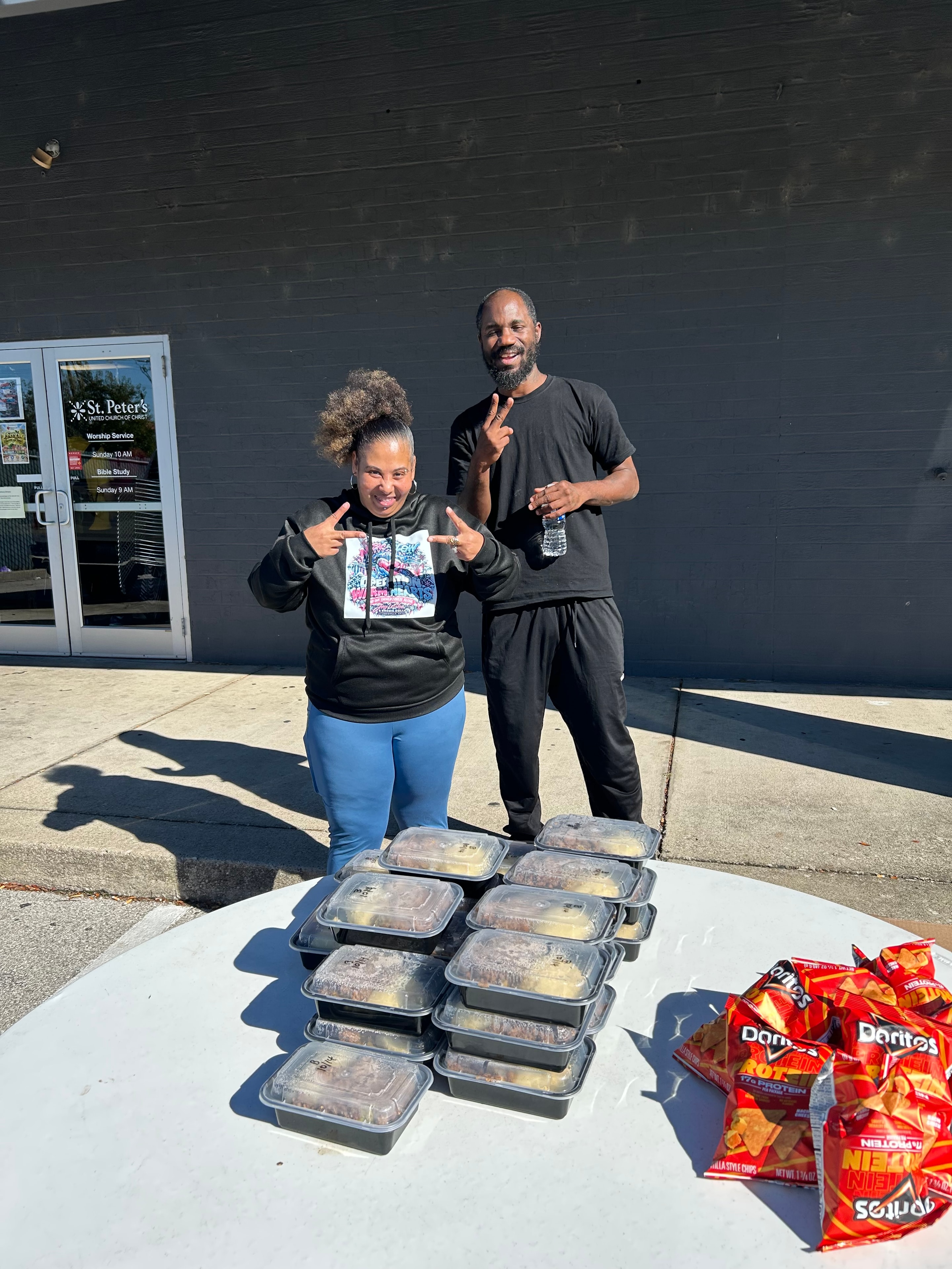 Two people posing with stacked hot meal containers and snacks ready for distribution